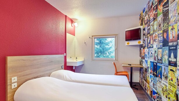A hotel room with a bed, a small desk with a TV, a window, and a wall decorated with travel posters. - hotelF1 Montpellier Sud (Rénové) (Lattes)