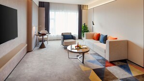 A modern hotel room with a sofa, armchair, coffee table, and a flat-screen TV.