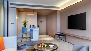 A modern hotel room with a sofa, a coffee table with fruit, a TV, and a small bar area.