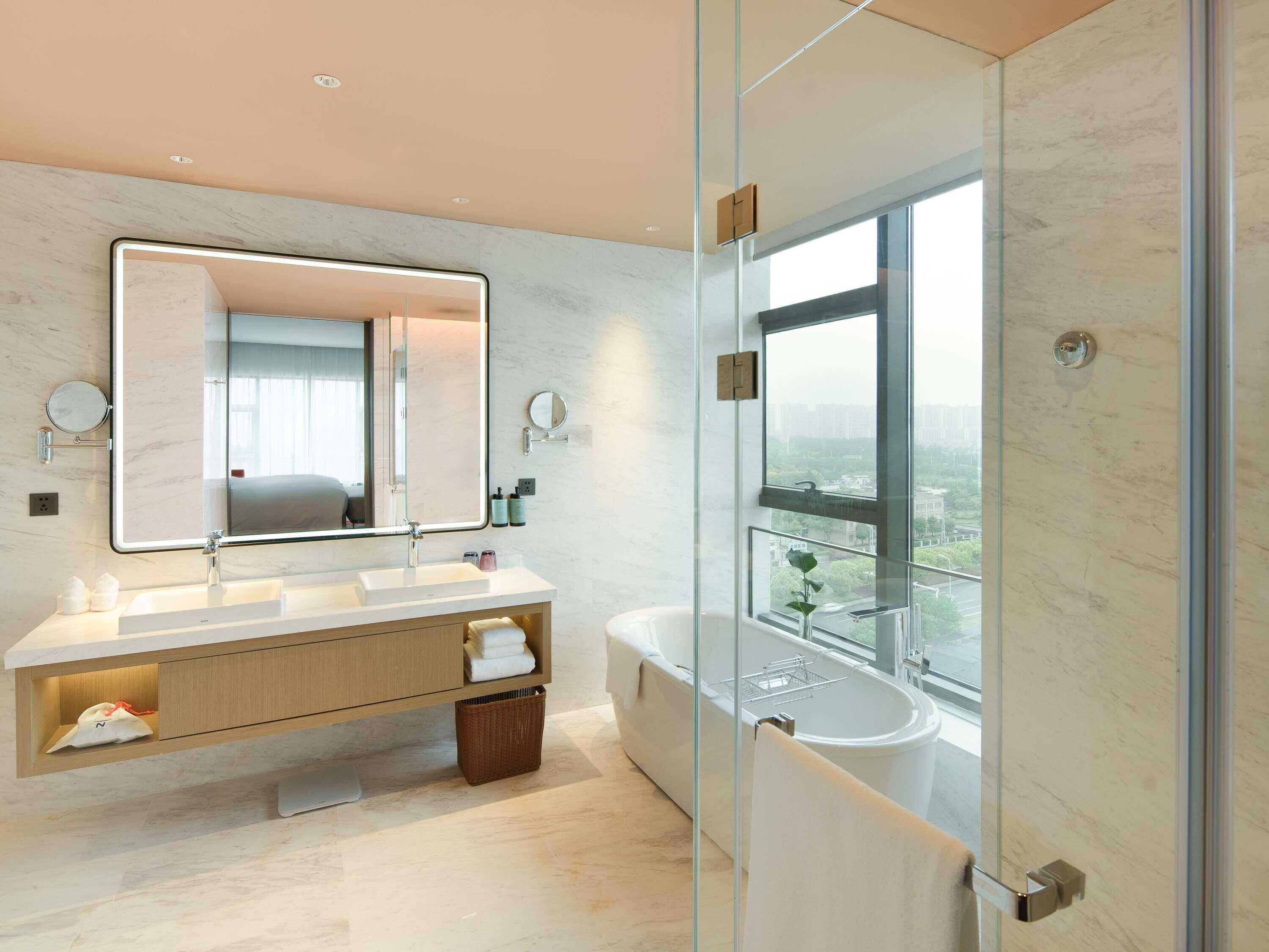 Un baño moderno con un espejo grande, lavamanos dobles, una bañera y vistas a través de una puerta de vidrio.
