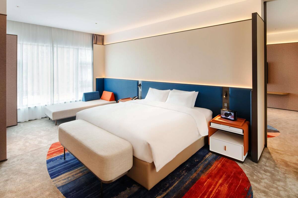 A modern hotel room with a large bed, bedside tables, a bench, and a view of the city through large windows.