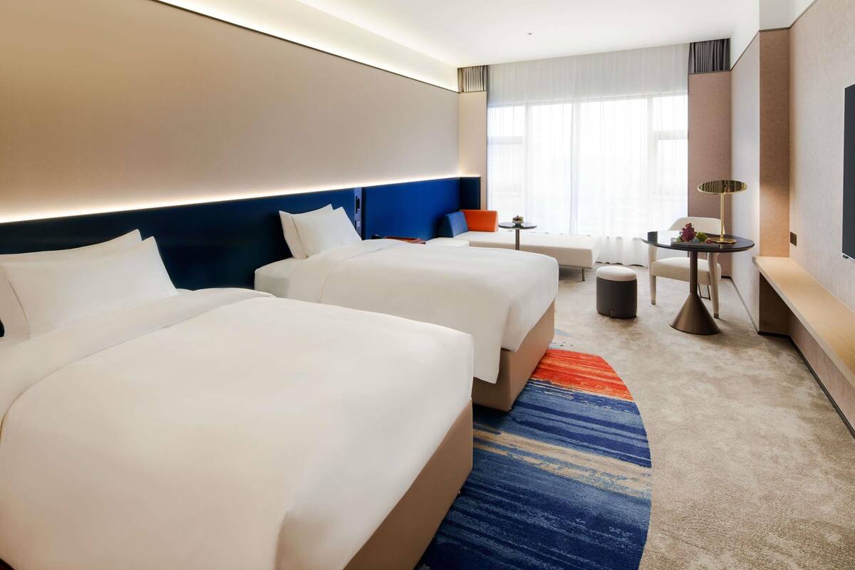 A hotel room with two beds, a small table, a bench, and a large window with curtains.