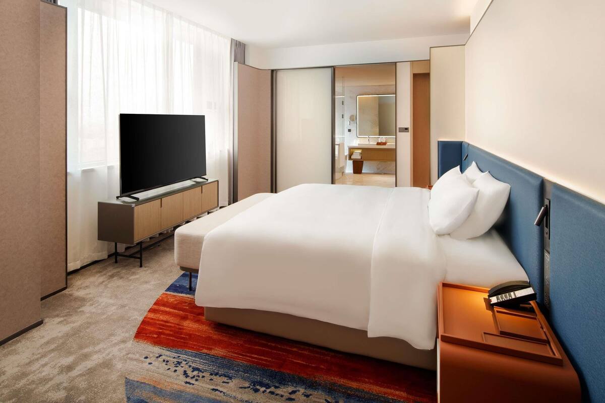 A hotel room with a bed, a TV, a desk, and a bathroom visible through an open door.