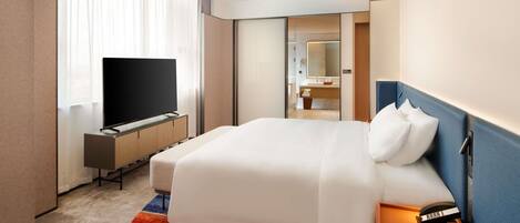Habitación de hotel con cama, televisor, escritorio y baño visible a través de una puerta abierta.