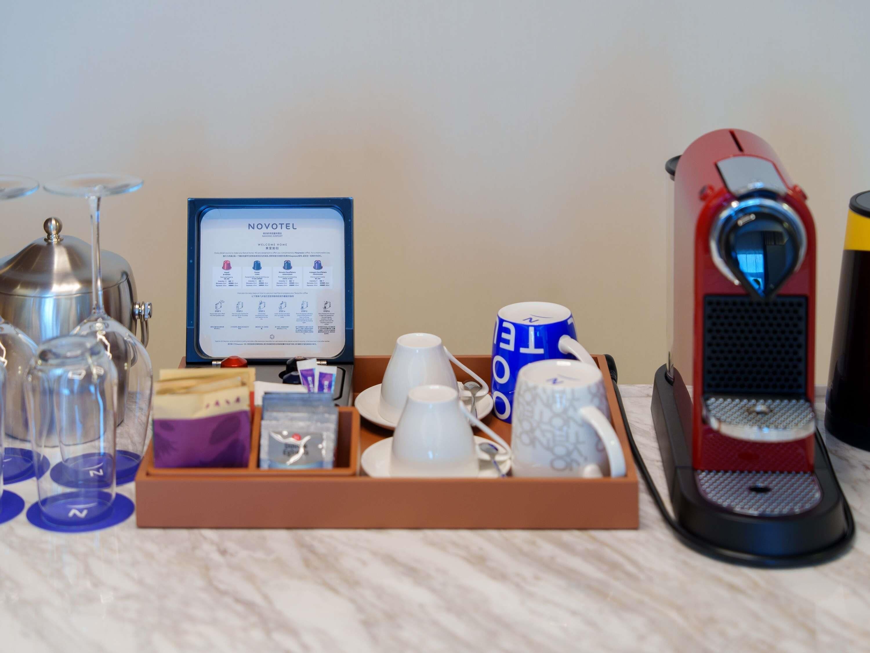 A hotel minibar setup with a coffee machine, tea and coffee mugs, a tea bag, a glass, and a menu board.
