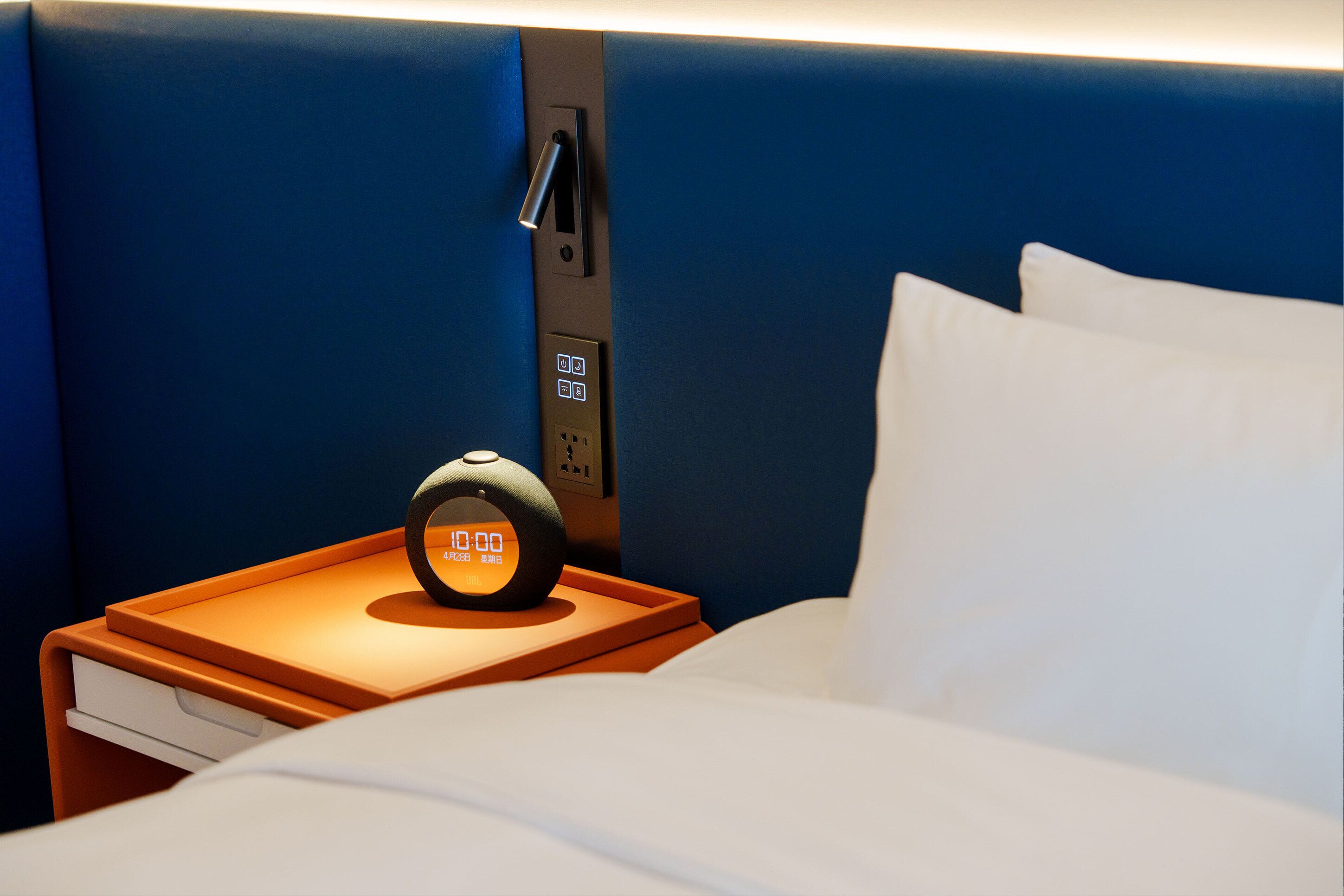 A hotel room with a bed, a nightstand with a digital clock, a power outlet, and a wall-mounted switch.