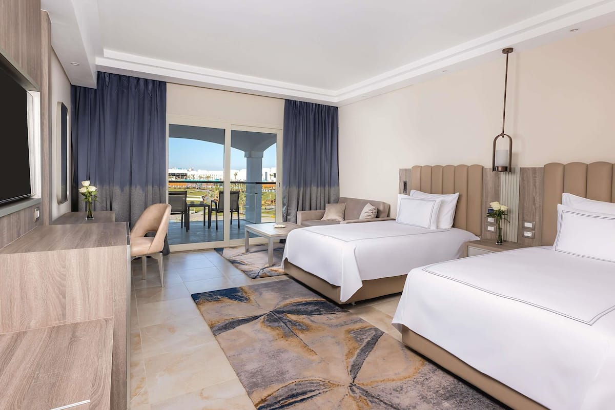 A hotel room with two beds, a TV, a desk, and a balcony with a view.