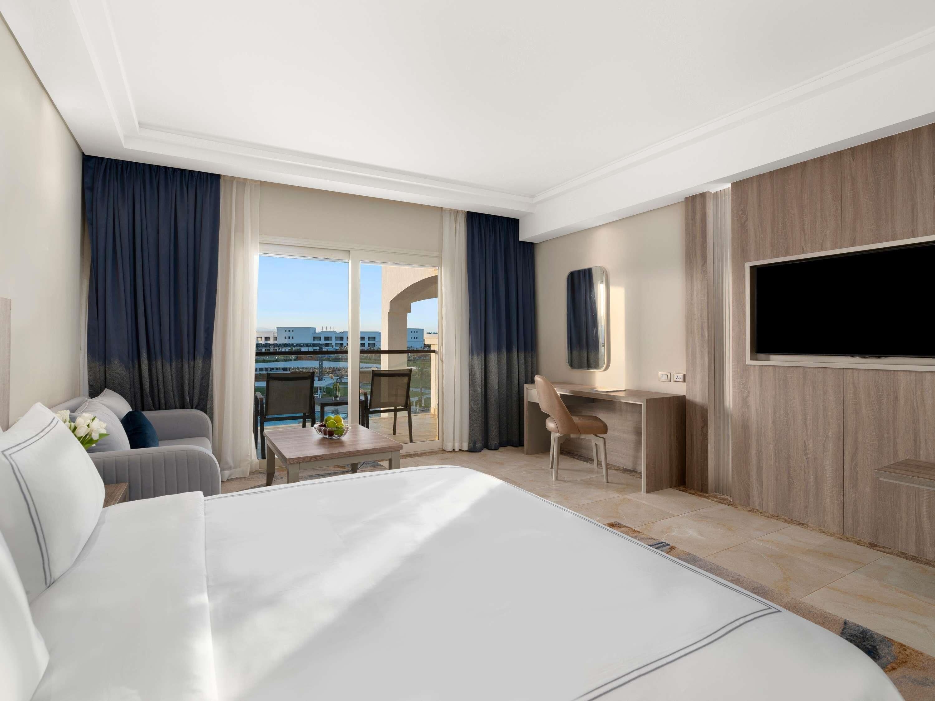 A hotel room with a bed, a desk, a chair, a TV, and a view of buildings through large windows.