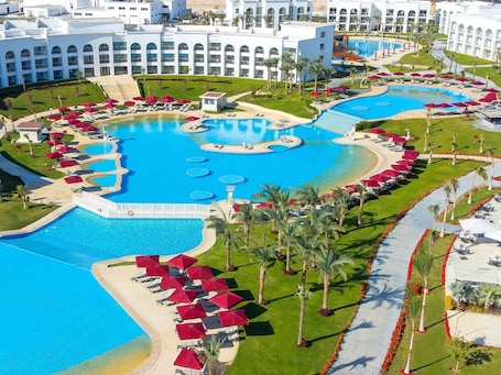 A large, multi-level swimming pool complex surrounded by palm trees and red umbrellas, with a white building in the background.