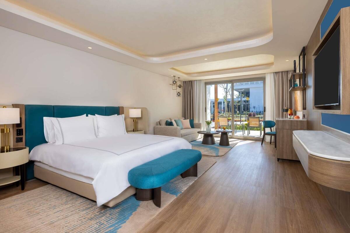 Una habitación de hotel con una cama grande, un sofá, una mesita, un banco, un televisor y un balcón con vistas.