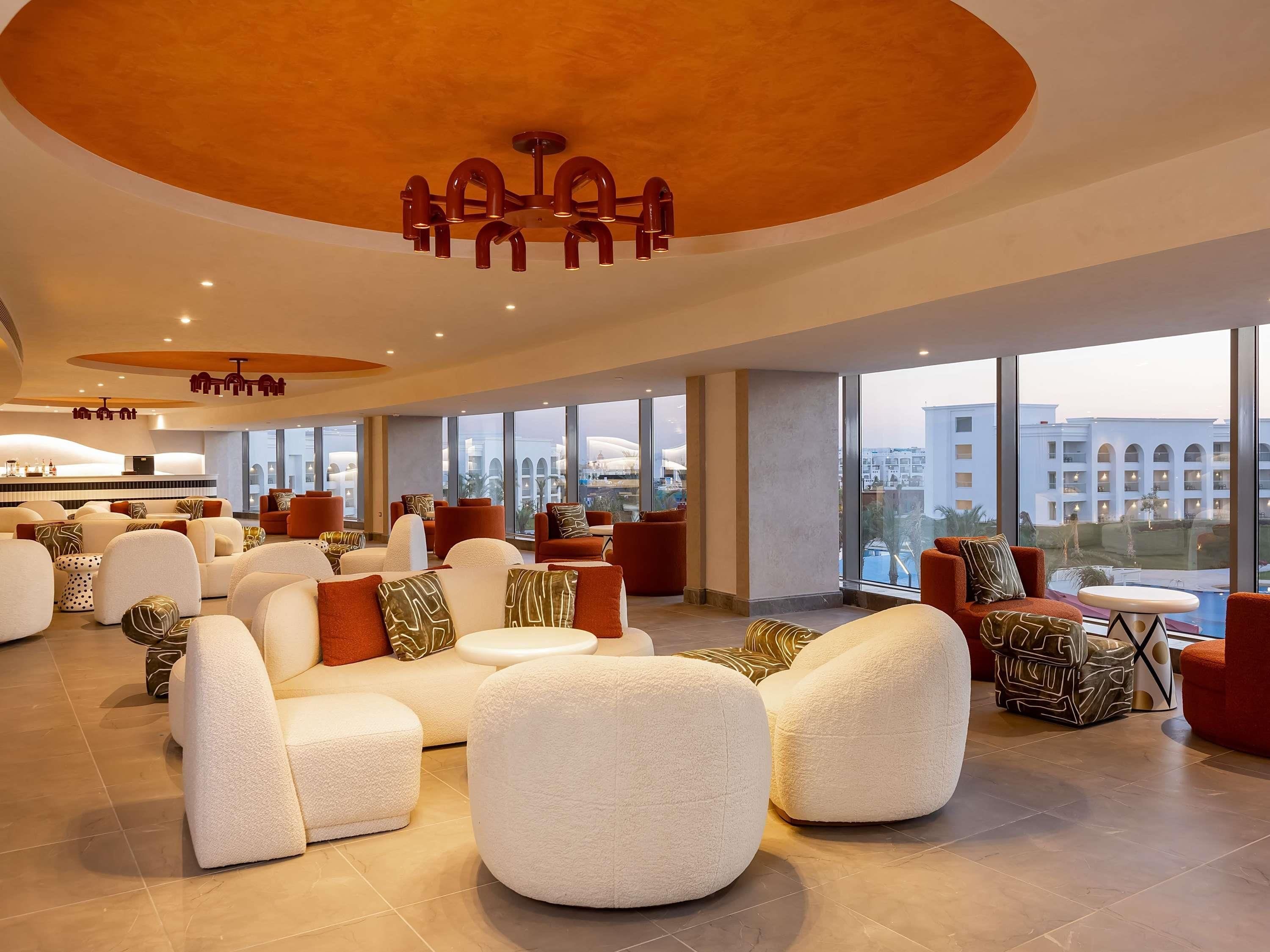 A spacious lobby with a modern design, featuring a central seating area with sofas and armchairs, a bar area, and large windows offering views of the surrounding buildings.