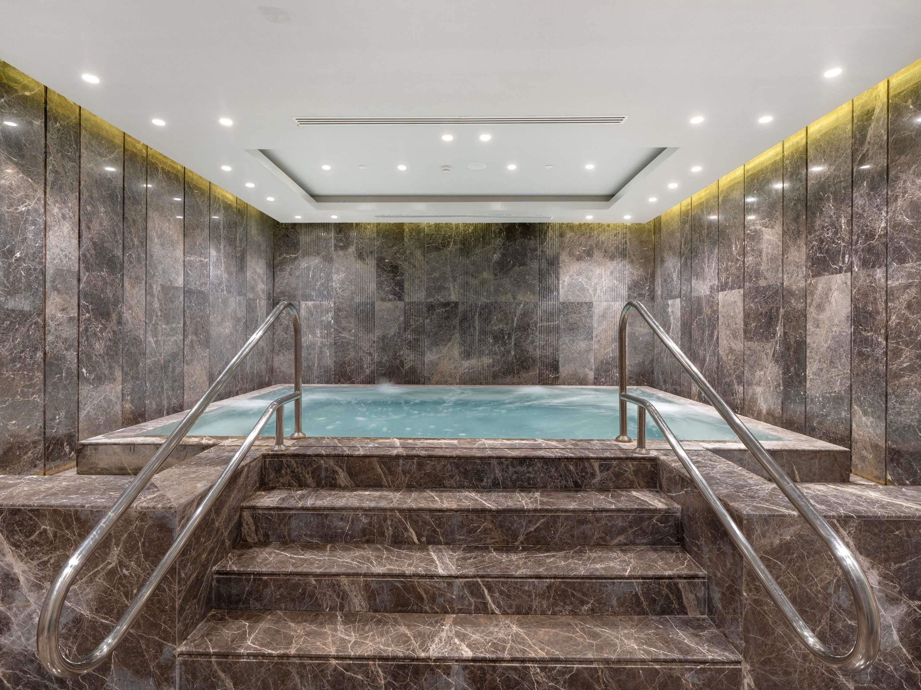 A marble-floored indoor pool with a central hot tub, surrounded by walls and a ceiling with recessed lighting.