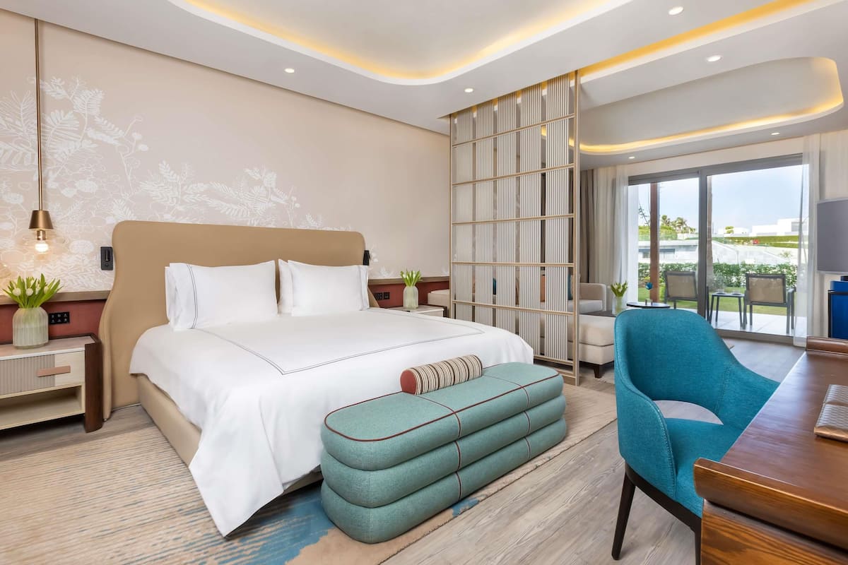 A modern hotel room with a large bed, a seating area with a table, a TV, and a view of the outdoors.