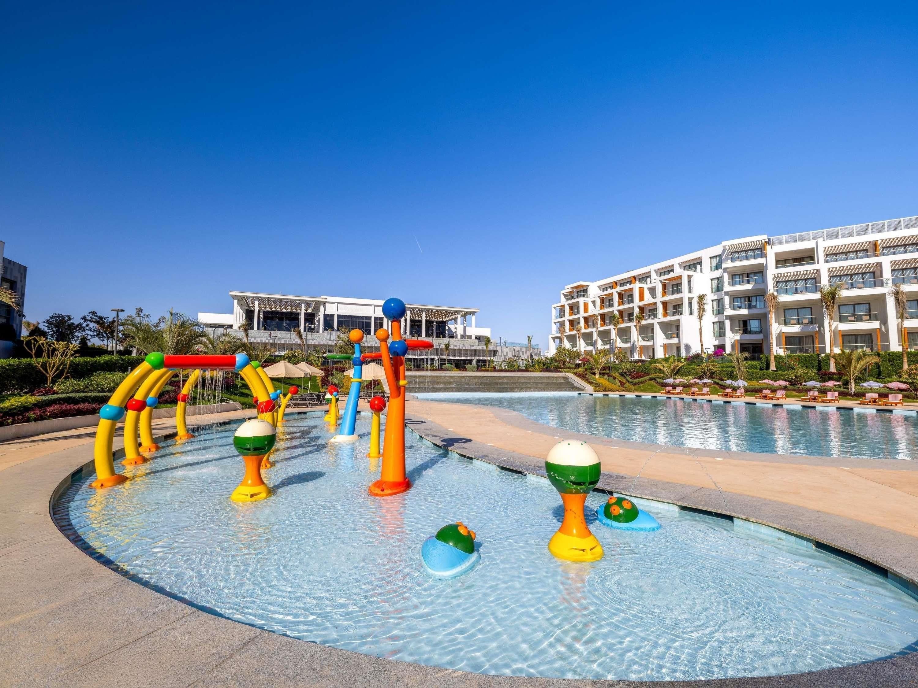 A swimming pool area with water play structures, a pool, and a hotel building in the background.