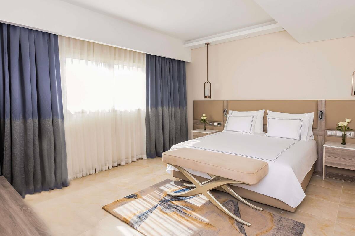 A hotel room with a bed, bedside tables, a nightstand, a lamp, and a window with curtains.