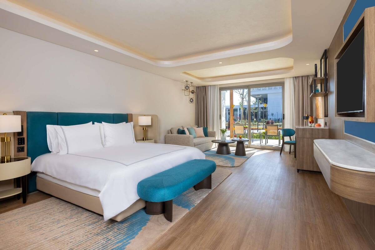 A modern hotel room with a large bed, a sofa, a small table, a bench, and a TV.