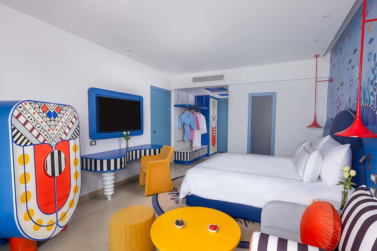 A modern hotel room with a large bed, a TV, a desk, a sofa, and a colorful wall decoration.