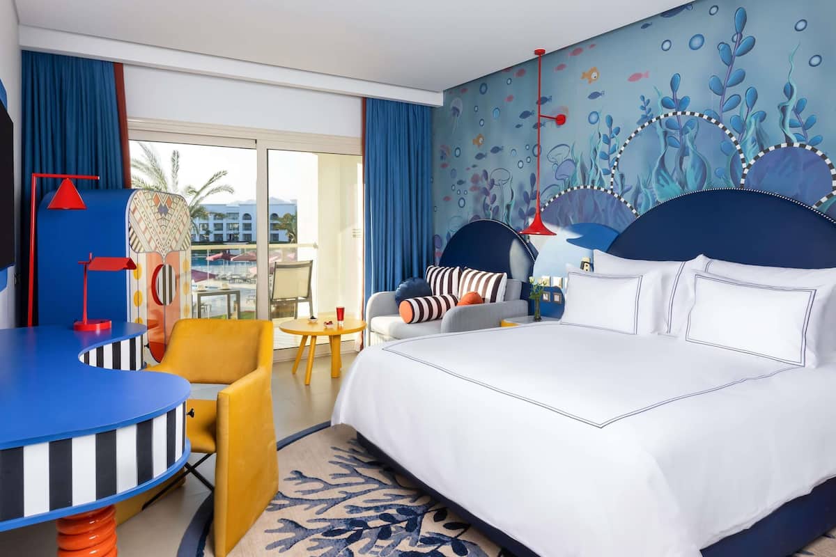 A hotel room with a bed, a desk, a chair, a small table, and a view of the outside.