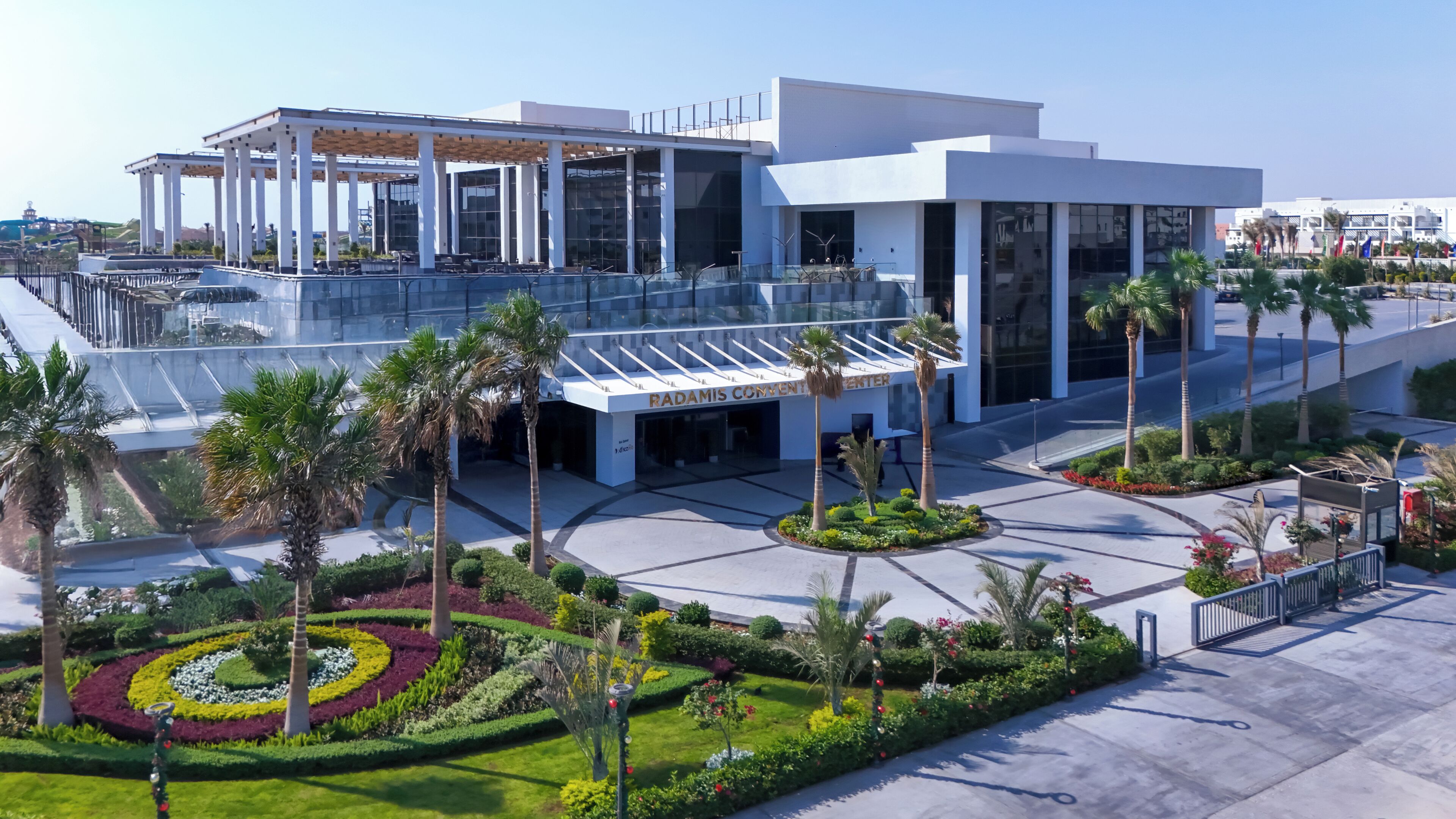 Un edificio moderno con fachada de vidrio, llamado "Radamis Convention Center", rodeado de jardines ajardinados y palmeras.