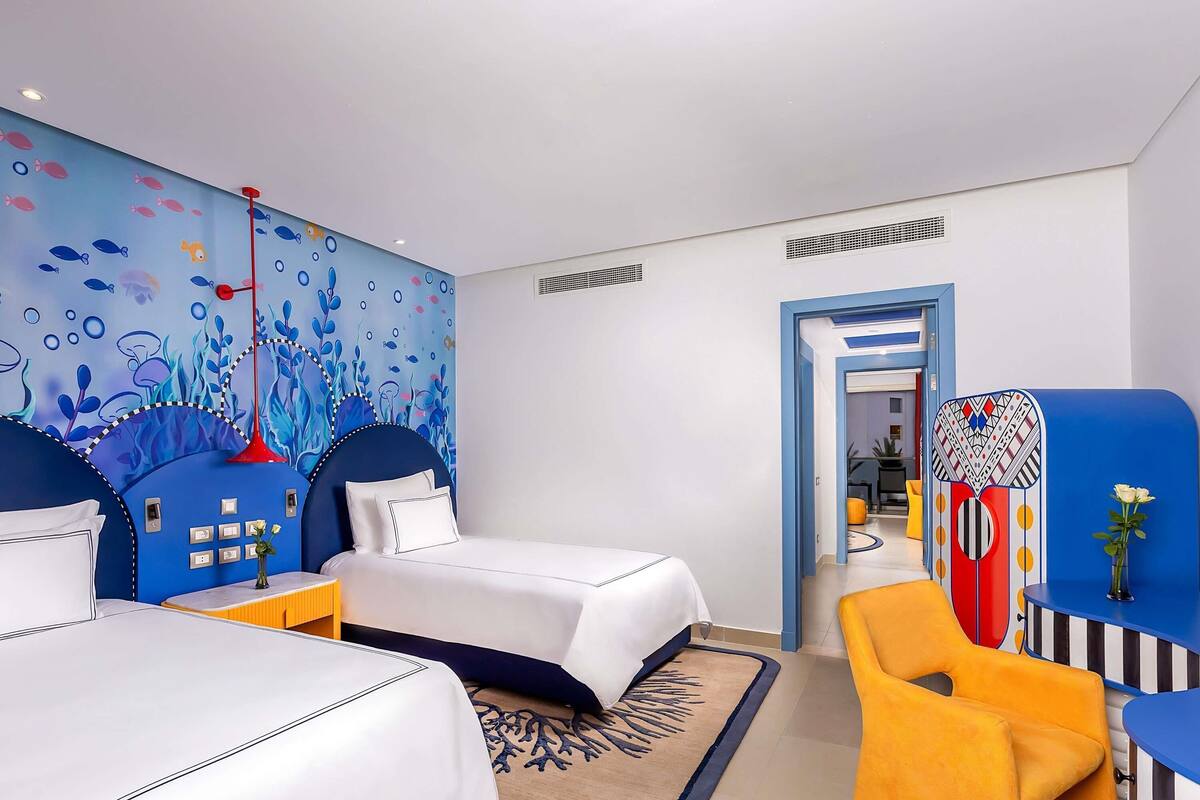 A hotel room with two beds, a vibrant mural, a yellow chair, and a view into the hallway.
