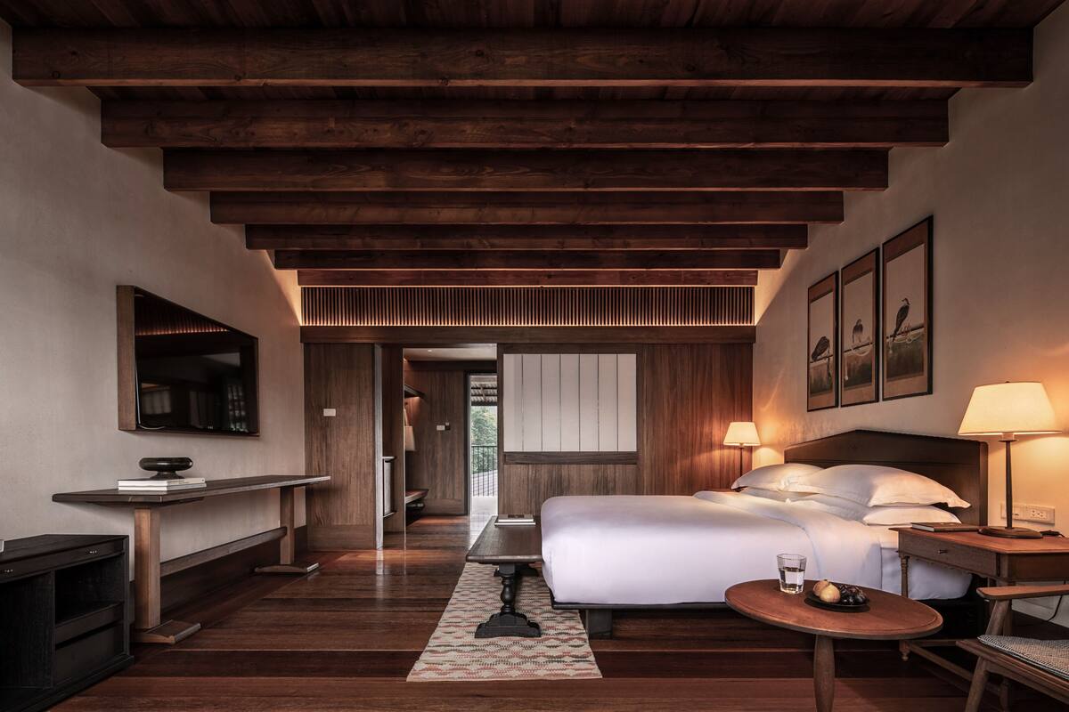 Une chambre d’hôtel avec un plafond en bois, un lit, une table de chevet avec une lampe, une petite table avec un verre d’eau et un téléviseur fixé au mur.