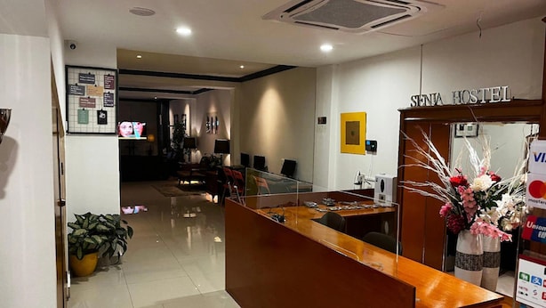 A reception area with a wooden desk, a reception counter, and a sign that reads "SENIA HOSTEL". There are potted plants and a flower arrangement on the counter. - Hotel O Senja Hostel Chinatown (Kuala Lumpur)