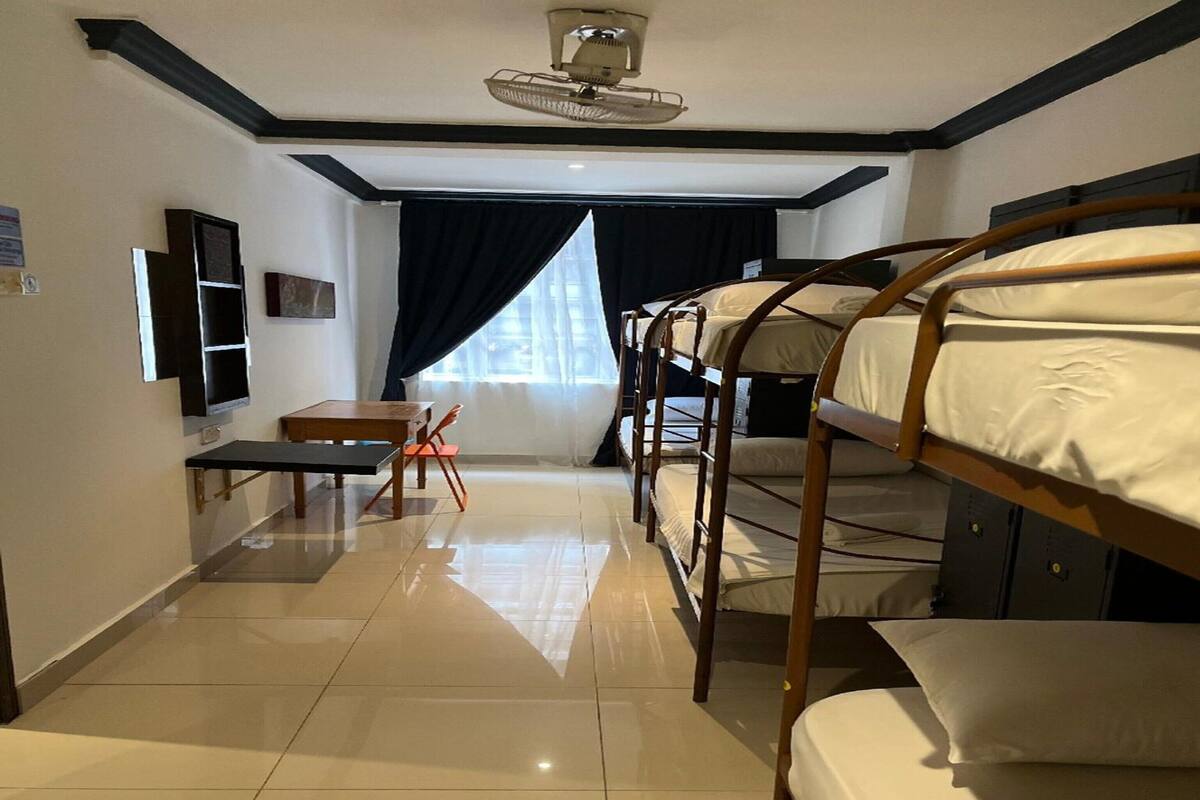 Hotelkamer met stapelbedden, een tafeltje, een stoel, een plafondventilator en een raam met gordijnen.