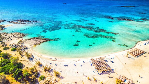Vista aérea de una playa con aguas turquesas cristalinas, una orilla de arena y varios sombrillas y tumbonas.
