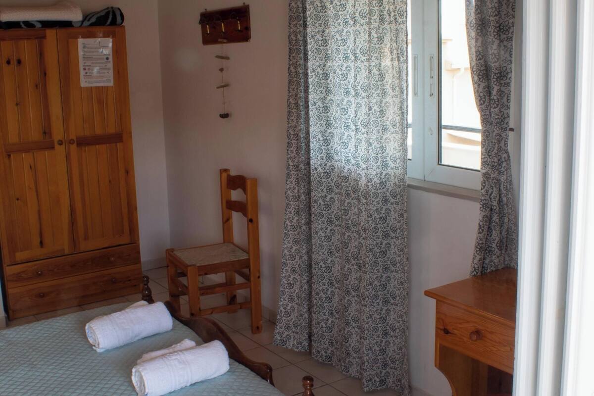 Un dormitorio con cama, armario de madera, silla, escritorio y ventana con cortinas estampadas.