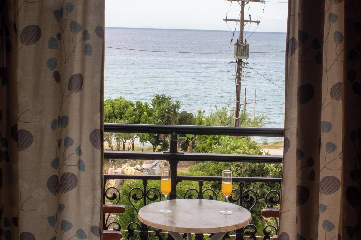 Un balcón con mesa y sillas, que ofrece vistas al mar con vegetación y una línea de electricidad.