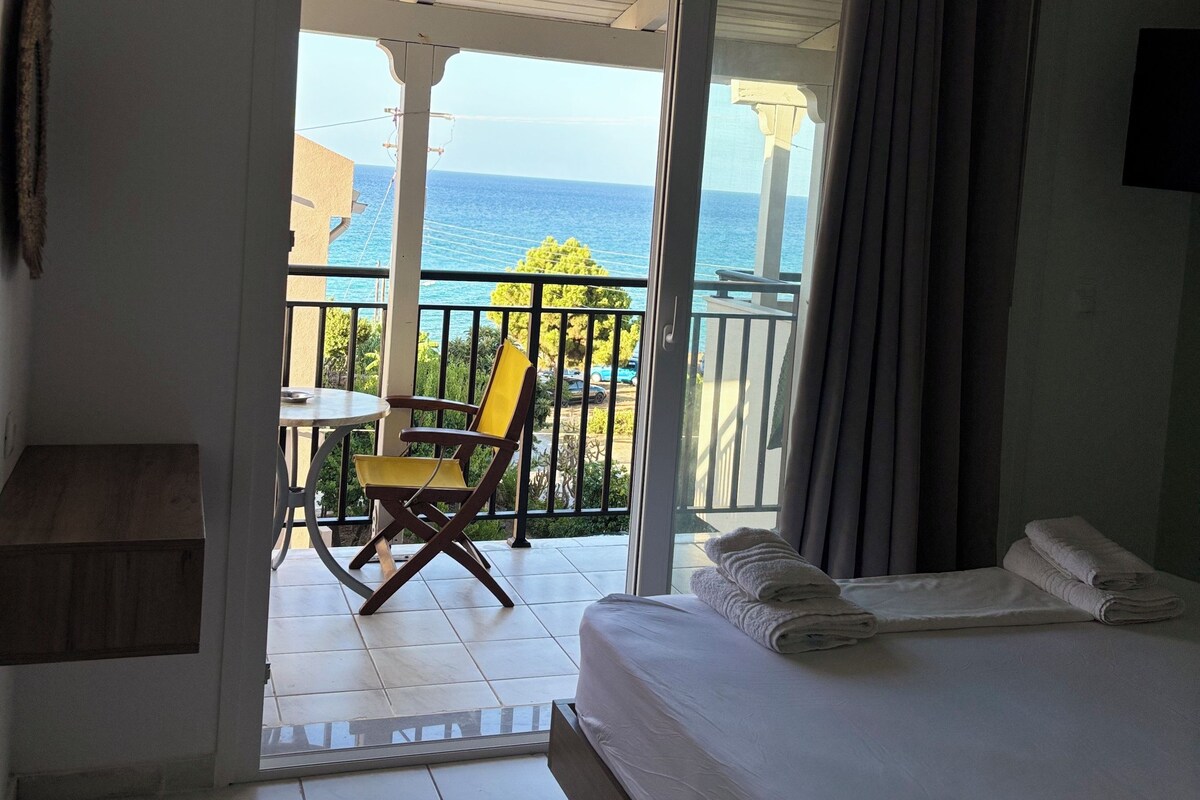 A hotel room with a bed, a small table, a chair, and a balcony with a view of the sea.