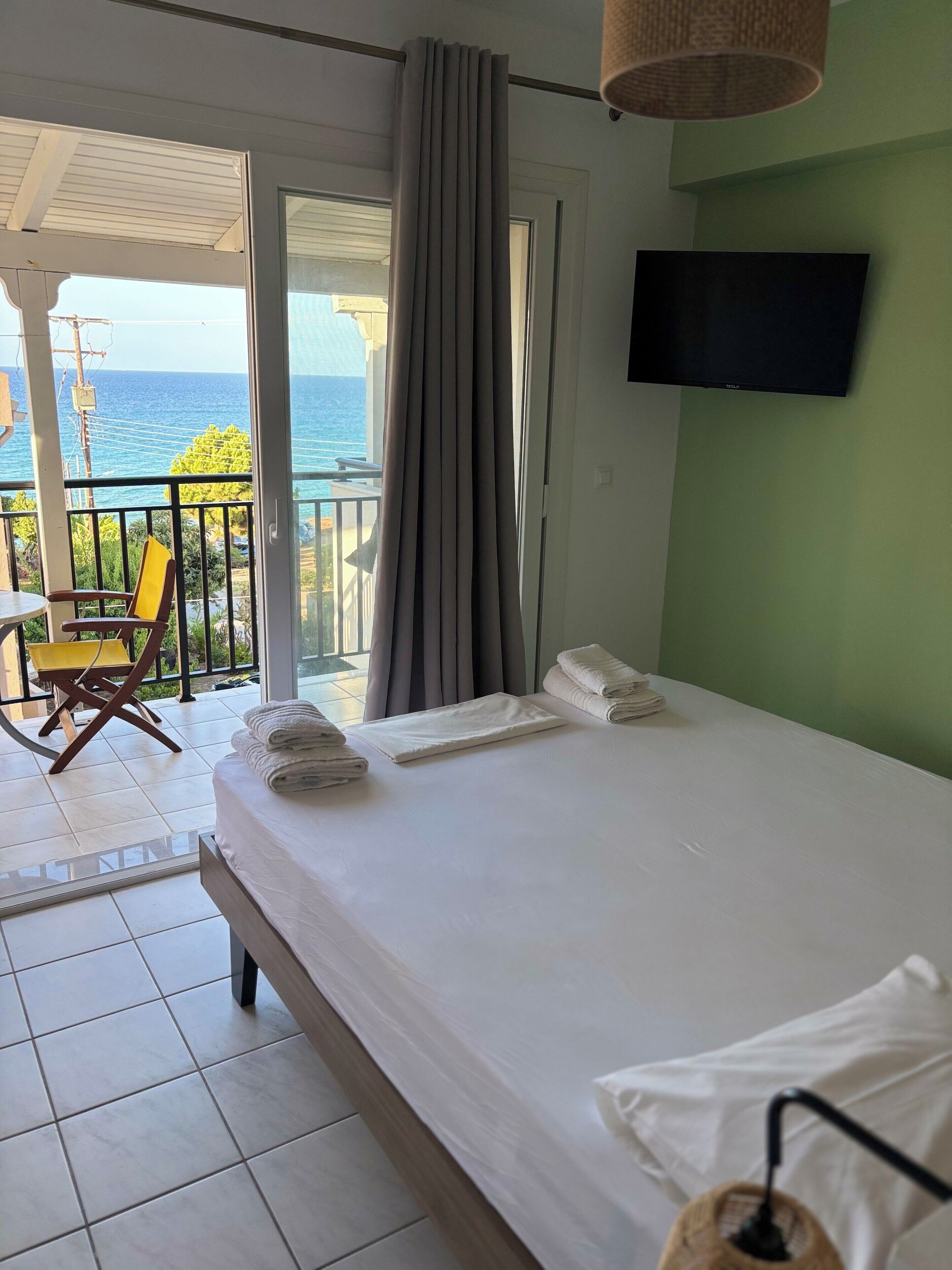 Ett hotellrum med en säng, en TV, en balkong med en stol och utsikt över havet.