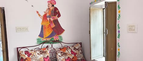 A bedroom with a floral bedspread, a decorative wall mural, a wooden door, and a window with a view.