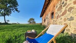 Zona verde con un edificio de piedra, una silla de descanso azul, una mesa pequeña con una taza y un libro.