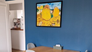 A dining area with a wooden table and four grey chairs, a blue wall with a framed painting, and a wooden floor. - Appartement Familial Proche de la Gare de Clamart (Clamart)