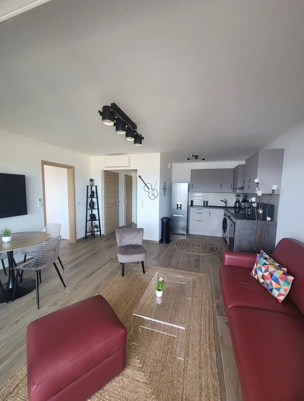 Een moderne woonkamer met een rode bank, een transparante salontafel en een grijze fauteuil. In de achtergrond is een keuken zichtbaar met grijze kasten en witte apparaten.