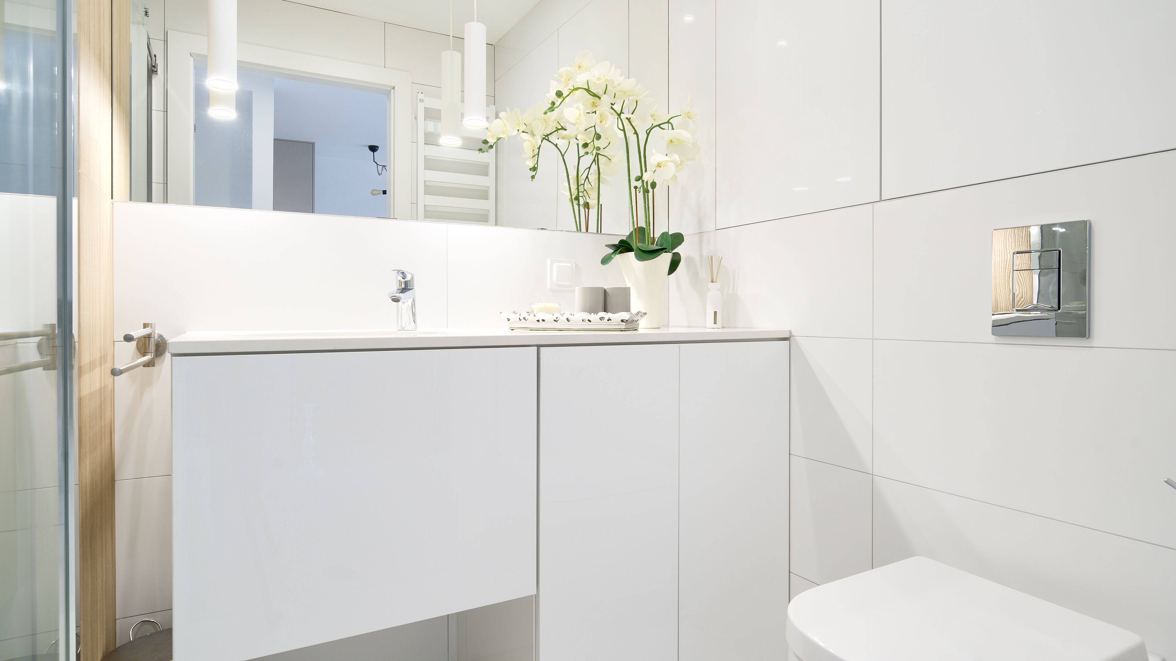 A modern bathroom with a white vanity, a mirror, and a toilet. There is a vase with white flowers on the vanity.