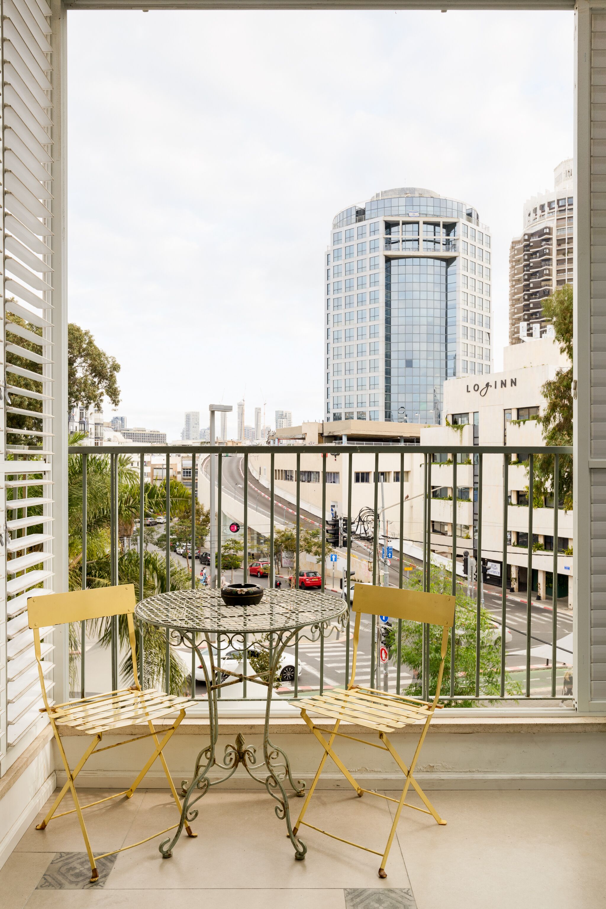 A balcony with a metal table and two chairs, overlooking a cityscape with tall buildings and a busy street.
