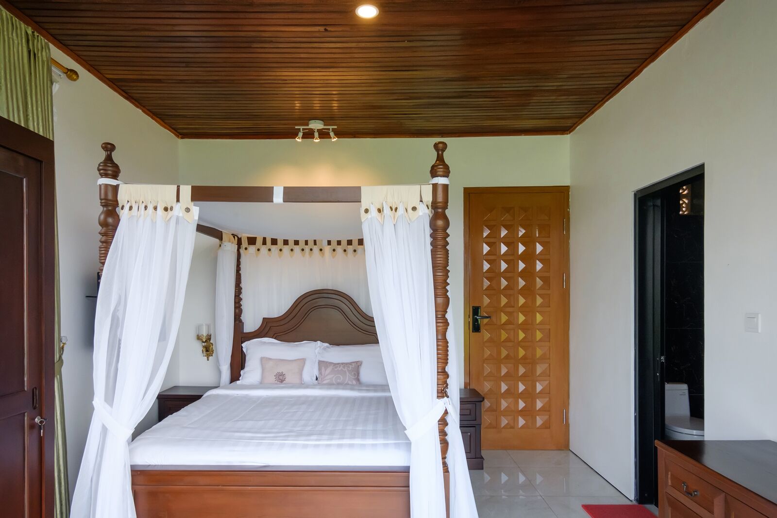 Un lit à baldaquin avec du linge de lit blanc, un plafond en bois et une porte en bois ornée d’un motif géométrique.