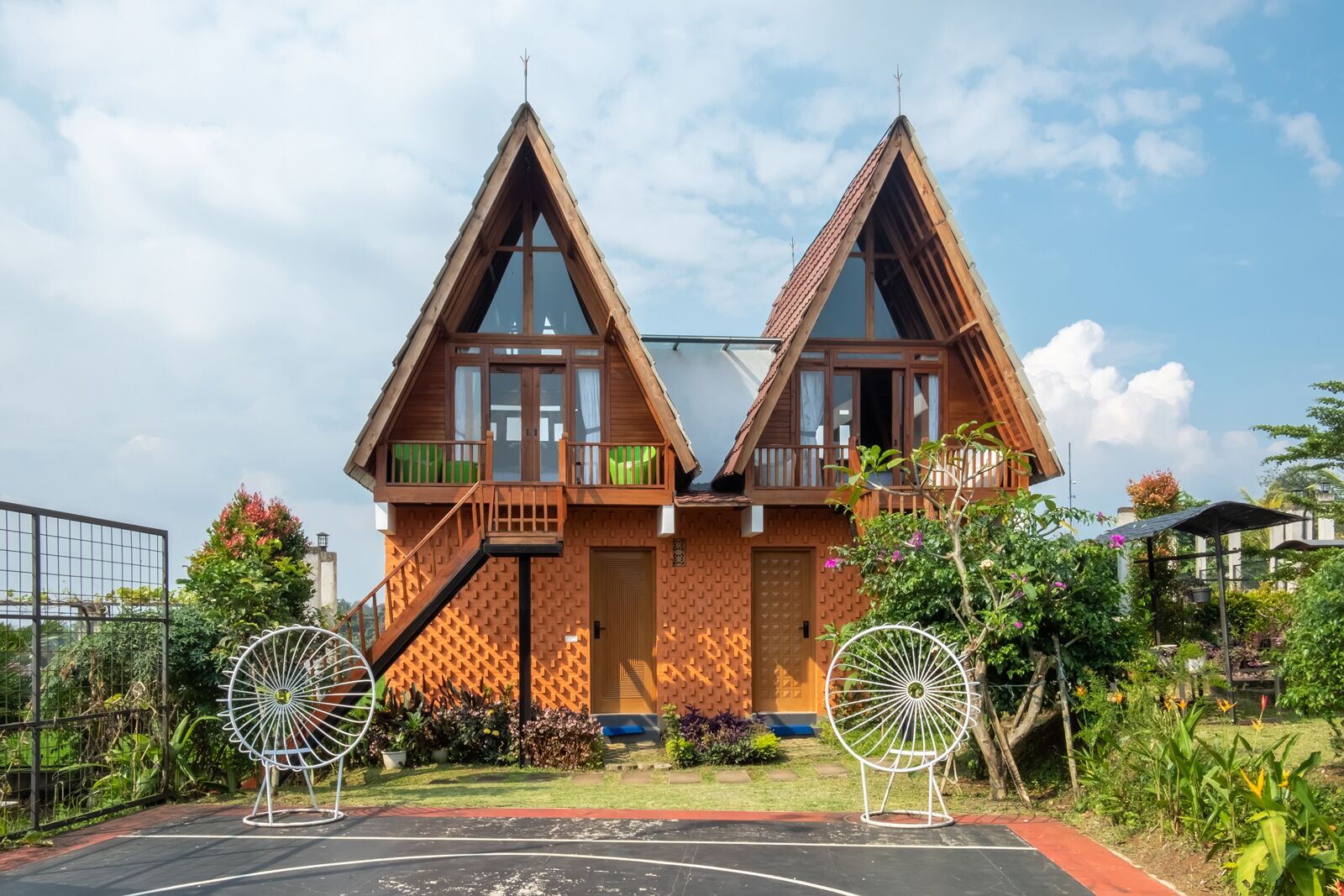 Een symmetrisch houten huis met twee balkons, een centrale deur en een kleine tuin met twee decoratieve windvaan-spinners.