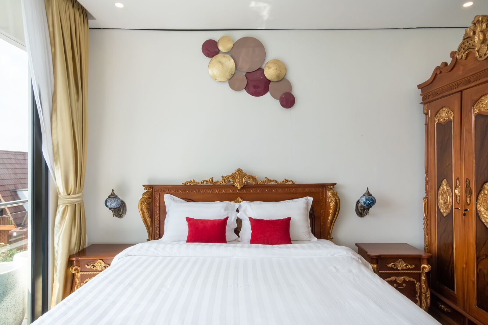 Habitación de hotel con una cama grande, cabecera de madera ornamentada, dos mesitas de noche, un armario y un adorno de pared.