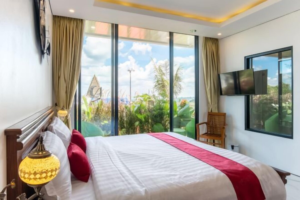 A hotel room with a large bed, a TV mounted on the wall, and a view of greenery outside.