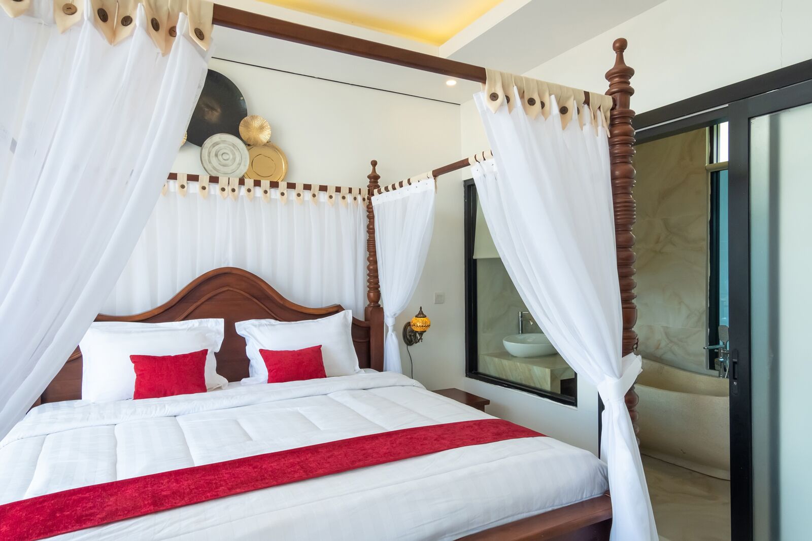A four-poster bed with white linens and red accents, a bedside table with a lamp, and a view of a bathroom through a glass door.