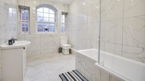 Un baño con bañera, inodoro y lavamanos, con azulejos de mármol y ventanas amplias.