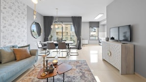 Ein modernes Wohnzimmer mit einer Couch, einem Esstisch, Stühlen, einem Fernseher und einem großen Fenster mit Vorhängen.