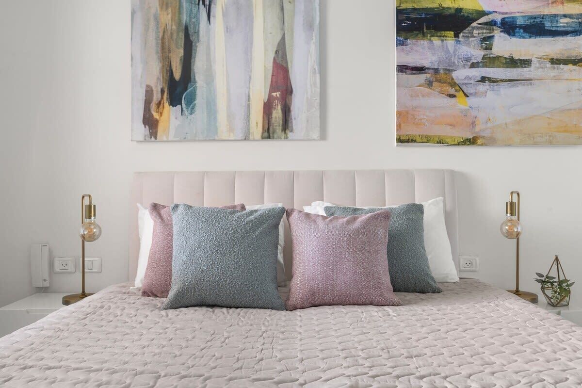 Une chambre à coucher avec une couvre-lit blanche matelassée, une tête de lit capitonnée et de nombreux coussins aux couleurs pastel.