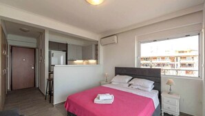 Une chambre à coucher avec un lit, des tables de chevet, une fenêtre et, au fond, une petite cuisine.