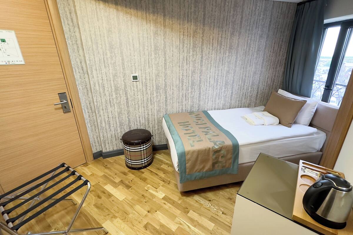 A compact hotel room with a bed, a small stool, a kettle, and a wooden floor.