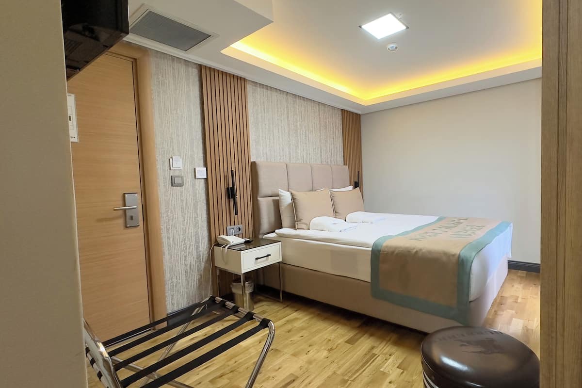 A modern hotel room with a wooden floor, a bed with a beige and blue bedspread, a bedside table with a phone, and a small stool.