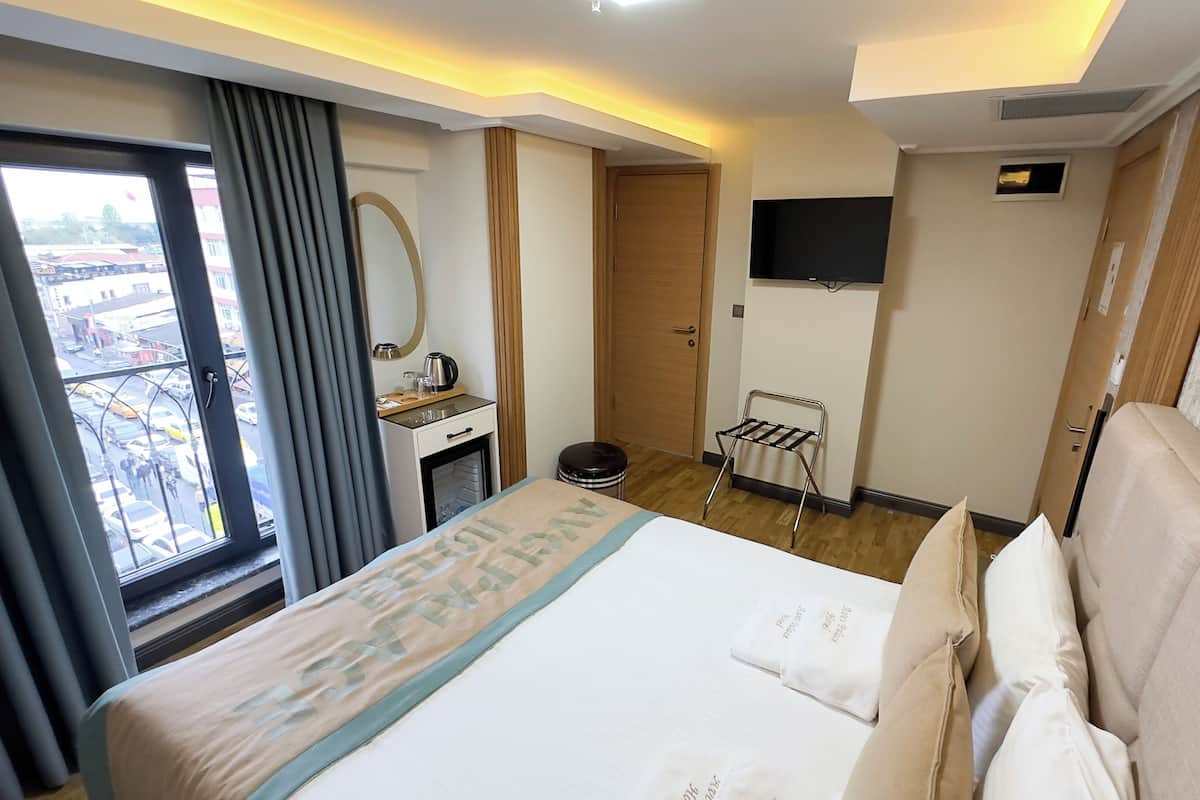 A hotel room with a bed, a desk with a mirror, a small window, a wall-mounted TV, and a view of the outside.